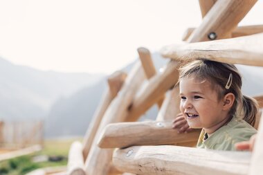 Lachendes Mädchen zwischen den Holzelementen eines Klettergerüsts auf dem Spielplatz am Schafberg in Gargellen. | © Gargellner Bergbahnen GmbH & Co KG, Daniel Zangerl