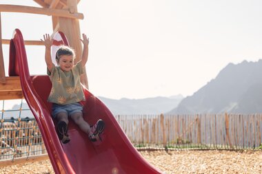 Kleines Mädchen rutscht auf dem Spielplatz am Schafberg eine rote Rutsche hinunter, im Hintergrund ist die Gargellner Bergwelt zu sehen. | © Gargellner Bergbahnen GmbH & Co KG, Daniel Zangerl