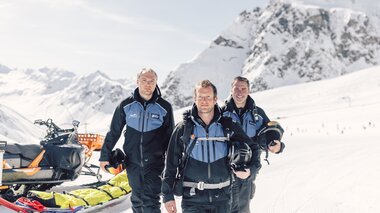 Drei Mitglieder der Pistenrettung Gargellen stehen gemeinsam im Schnee, bereit für ihren nächsten Einsatz. Hinter ihnen ein Schneemobil mit Rettungsschlitten, im Hintergrund die verschneiten Gipfel des Montafons. Ihre Entschlossenheit und Professionalität prägen das Bild – Sicherheit steht an erster Stelle. | © Gargellner Bergbahnen GmbH & Co KG, Daniel Zangerl
