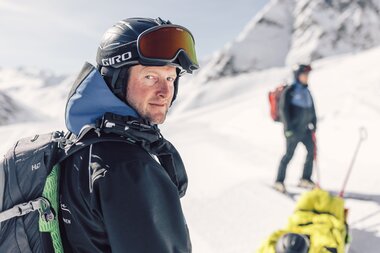 Ein Mitglied der Pistenrettung in Gargellen blickt konzentriert in die Kamera – bereit für den nächsten Einsatz im alpinen Gelände. Im Hintergrund kümmern sich Kolleg:innen um einen verletzten Wintersportler, während die Sonne auf den verschneiten Hang scheint. | © Gargellner Bergbahnen GmbH & Co KG, Daniel Zangerl