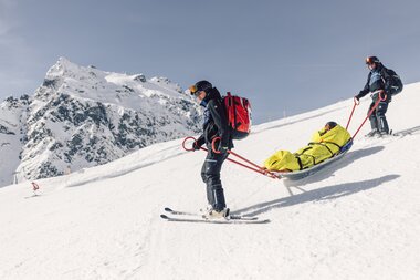Zwei Mitglieder der Pistenrettung bringen einen verletzten Skifahrer im Akia sicher ins Tal. Mit großer Routine steuern sie den Akia durch das verschneite Skigebiet von Gargellen, um eine schnelle und sichere Versorgung zu gewährleisten. | © Gargellner Bergbahnen GmbH & Co KG, Daniel Zangerl
