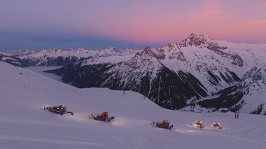 Mehrere Pistenraupen der Gargellner Bergbahnen präparieren bei Sonnenuntergang die schneebedeckten Hänge des Skigebiets. Der Himmel leuchtet in zarten Rosa- und Lilatönen, während sich die umliegenden Berge majestätisch im Hintergrund erheben. Eine friedliche Winterstimmung in der hochalpinen Landschaft von Gargellen. | © Montafon Tourismus GmbH, Marie Schilcher