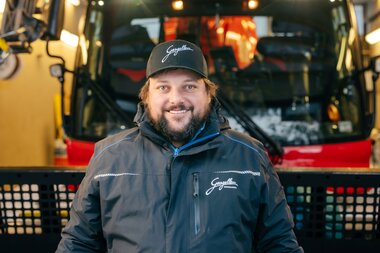 Ein Mitarbeiter der Gargellner Bergbahnen steht lächelnd in der Werkstatt vor einer roten Pistenraupe. Er trägt eine dunkelblaue Jacke und eine schwarze Kappe mit dem Schriftzug „Gargellen“. Im Hintergrund ist die beleuchtete Fahrzeughalle mit der Pistenraupe zu sehen. | © Montafon Tourismus GmbH, Marie Schilcher