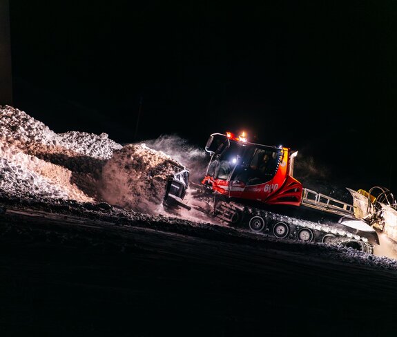 Eine rote Pistenraupe der Gargellner Bergbahnen schiebt in der Nacht frischen Schnee über die Piste. Im Schein der starken Scheinwerfer wirbelt der Schnee auf und glitzert im Licht – ein eindrucksvolles Bild der nächtlichen Präparationsarbeiten im Skigebiet Gargellen. | © Montafon Tourismus GmbH, Marie Schilcher