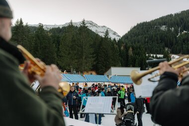Blick von der Bühne über die Schulter zweier Musiker mit Blasinstrumenten auf zahlreiche Besucher eines Wintermarkts; Marktstände mit blau-weißen Dächern stehen vor einem bewaldeten Berghang | © Montafon Tourismus, Marie Schilcher
