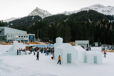 Wintermarkt in Gargellen mit Besucherinnen und Besuchern auf einer verschneiten Fläche, großen Schnee- und Eisbauwerken im Vordergrund sowie Marktständen und Bergbahnen vor bewaldeten Hängen und verschneiten Gipfeln. | © Montafon Tourismus, Marie Schilcher 