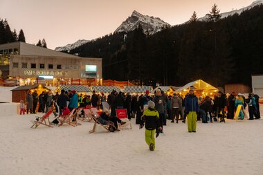 Belebter Wintermarkt in Gargellen mit beleuchteten Marktständen, Gästen im Schnee, Stehtischen und Liegestühlen vor verschneiten Bergen und dunklem Nadelwald in der Abenddämmerung. | © Montafon Tourismus, Marie Schilcher