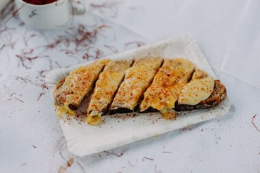 Eine Scheibe dunkles Brot ist mit geschmolzenem Käse überbacken und in Streifen geschnitten; das Brot liegt auf Papier, darüber ist Paprikapulver gestreut, im Hintergrund sind Safranfäden und eine Tasse zu sehen. | © Montafon Tourismus, Marie Schilcher