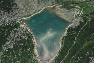 Ein faszinierendes Luftbild zeigt den türkisblauen Gandasee in Gargellen, eingebettet zwischen Felsen, Wiesen und Bergpfaden. Das klare Wasser spiegelt die Wolken am Himmel – ein Ort der Ruhe und Ursprünglichkeit inmitten der Montafoner Bergwelt. | © Gargellner Bergbahnen GmbH & Co KG, Daniel Zangerl