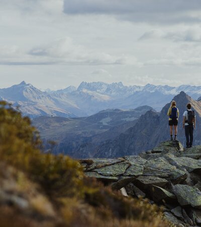Zwei Wandernde stehen auf einem Felsen hoch über Gargellen und genießen den Blick auf die umliegenden Berge. Der Herbst taucht die Landschaft in sanfte Farben, während die schneebedeckten Gipfel in der Ferne leuchten – ein Moment der Ruhe und Weite in Gargellen. | © Gargellner Bergbahnen GmbH & Co KG, Alexander Furchs 