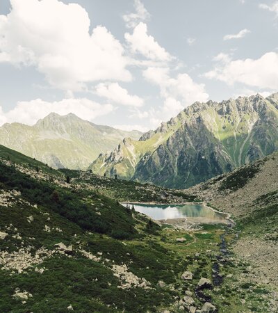 Der Gandasee liegt ruhig eingebettet zwischen steilen Felswänden und grünen Hängen oberhalb von Gargellen. Im klaren Wasser spiegeln sich Wolken und Gipfel, während ein kleiner Bach aus dem Tal heraufplätschert. Eine eindrucksvolle Szenerie, die die alpine Ursprünglichkeit des Montafons widerspiegelt. | © Gargellner Bergbahnen GmbH & Co KG, Daniel Zangerl