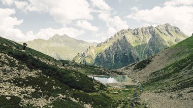 Der Gandasee liegt ruhig eingebettet zwischen steilen Felswänden und grünen Hängen oberhalb von Gargellen. Im klaren Wasser spiegeln sich Wolken und Gipfel, während ein kleiner Bach aus dem Tal heraufplätschert. Eine eindrucksvolle Szenerie, die die alpine Ursprünglichkeit des Montafons widerspiegelt. | © Gargellner Bergbahnen GmbH & Co KG, Daniel Zangerl