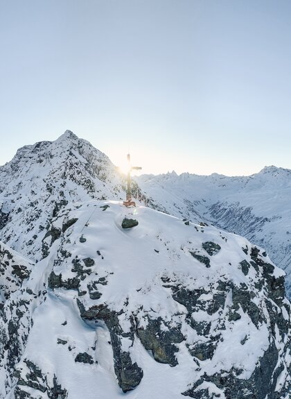 Verschneiter Berggipfel mit Gipfelkreuz im Sonnenlicht, umgeben von einer beeindruckenden winterlichen Berglandschaft im Montafon. | © Gargellner Bergbahnen GmbH & Co KG, Daniel Zangerl