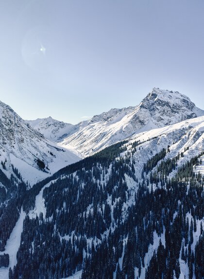 Panoramablick auf die verschneiten Gipfel und Berghänge rund um Gargellen. Die Sonne steht über einem Berggipfel, während das Bergdorf im Tal zwischen schneebedeckten Wäldern liegt. | © Gargellner Bergbahnen GmbH & Co KG, Daniel Zangerl