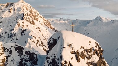 Gipfelkreuz auf einem verschneiten Berggipfel bei Gargellen im warmen Licht der Abendsonne. | © Gargellner Bergbahnen GmbH & Co KG, Daniel Zangerl