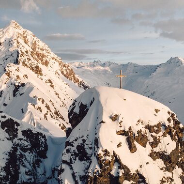 Gipfelkreuz auf einem verschneiten Berggipfel bei Gargellen im warmen Licht der Abendsonne. | © Gargellner Bergbahnen GmbH & Co KG, Daniel Zangerl