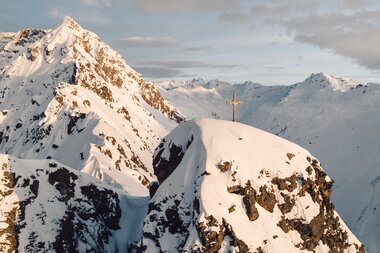 Gipfelkreuz auf einem verschneiten Berggipfel bei Gargellen im warmen Licht der Abendsonne. | © Gargellner Bergbahnen GmbH & Co KG, Daniel Zangerl