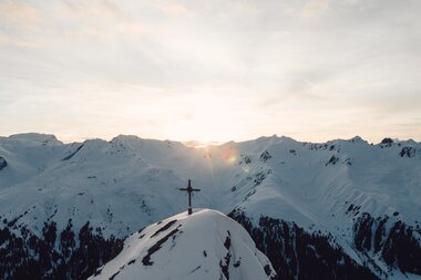 Die Sonne geht über den verschneiten Gipfeln von Gargellen auf und taucht das Gipfelkreuz in sanftes Licht. Ein Augenblick der Ruhe und Kraft, mitten in der winterlichen Bergwelt. | © Gargellner Bergbahnen GmbH & Co KG, Daniel Zangerl