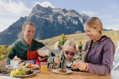 Eine Familie genießt ihr Frühstück auf einer Sonnenterrasse in den Bergen von Gargellen. Die Eltern und zwei Kinder sitzen an einem Holztisch mit Saft, Obst und Frühstückstellern. Im Hintergrund erheben sich grüne Berghänge und eine markante Felswand unter blauem Himmel. | © Gargellner Bergbahnen GmbH & Co KG, Alexander Fuchs