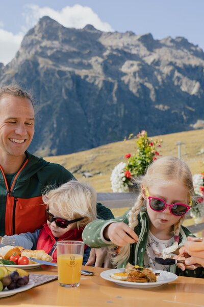 Eine Familie genießt ihr Frühstück auf einer Sonnenterrasse in den Bergen von Gargellen. Die Eltern und zwei Kinder sitzen an einem Holztisch mit Saft, Obst und Frühstückstellern. Im Hintergrund erheben sich grüne Berghänge und eine markante Felswand unter blauem Himmel. | © Gargellner Bergbahnen GmbH & Co KG, Alexander Fuchs