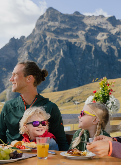 Eine Familie frühstückt auf einer Sonnenterrasse mit Blick auf die Berge von Gargellen. Eltern und Kinder sitzen an einem Holztisch, essen und lachen gemeinsam. Im Hintergrund ragt ein markanter Felsgipfel in den blauen Himmel, während andere Gäste auf der Terrasse sitzen. | © Gargellner Bergbahnen GmbH & Co KG, Alexander Fuchs