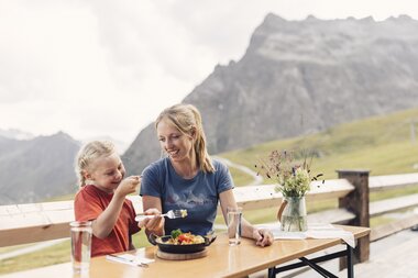 Eine Frau und ein Kind sitzen an einem Holztisch auf einer Sonnenterrasse und genießen gemeinsam ein Mittagessen mit Blick auf die grünen Berge von Gargellen. Beide lächeln, während sie eine Mahlzeit teilen. Auf dem Tisch stehen ein Glas Wasser und ein Krug mit Wiesenblumen. | © Gargellner Bergbahnen GmbH & Co KG, Daniel Zangerl
