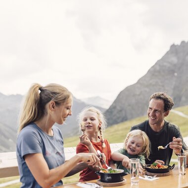 Eine Familie mit zwei Kindern sitzt an einem Holztisch auf einer Sonnenterrasse und genießt ihr Mittagessen mit Blick auf die Berge von Gargellen. Alle lachen und wirken entspannt. Auf dem Tisch stehen Teller mit bunten Gerichten, Gläser mit Wasser und eine Vase mit frischen Wiesenblumen. | © Gargellner Bergbahnen GmbH & Co KG, Daniel Zangerl