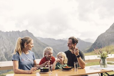 Eine Familie mit zwei Kindern sitzt an einem Holztisch auf einer Sonnenterrasse und isst gemeinsam mit Blick auf die Berge von Gargellen. Die Eltern und Kinder lachen und wirken entspannt. Auf dem Tisch stehen Teller mit buntem Essen, Gläser mit Wasser und eine Vase mit frischen Wiesenblumen. | © Gargellner Bergbahnen GmbH & Co KG, Daniel Zangerl