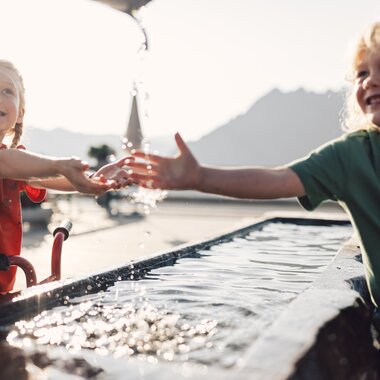 Zwei Kinder spielen fröhlich an einem Brunnen in der Sonne. Ein Mädchen mit rotem T-Shirt hält die Hände unter den Wasserstrahl, während ein Junge im grünen T-Shirt lachend daneben steht. Das Sonnenlicht spiegelt sich im Wasser, und im Hintergrund sind Berge leicht unscharf zu erkennen. | © Gargellner Bergbahnen GmbH & Co KG, Daniel Zangerl