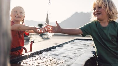 Zwei Kinder spielen fröhlich an einem Brunnen in der Sonne. Ein Mädchen mit rotem T-Shirt hält die Hände unter den Wasserstrahl, während ein Junge im grünen T-Shirt lachend daneben steht. Das Sonnenlicht spiegelt sich im Wasser, und im Hintergrund sind Berge leicht unscharf zu erkennen. | © Gargellner Bergbahnen GmbH & Co KG, Daniel Zangerl