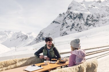 Ein Mann und eine Frau sitzen an einem Tisch auf der Terrasse eines Bergrestaurants und genießen jeweils ein Essen und ein warmes Getränk.  | © Gargellner Bergbahnen GmbH & Co KG, Daniel Zangerl