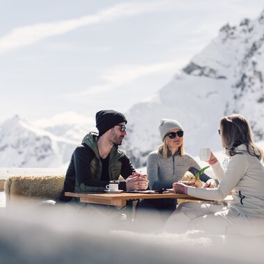 Drei Personen sitzen auf einer mit Fellen belegten Bank an einem Holztisch auf einer Sonnenterrasse in den Bergen. Sie tragen Winterkleidung mit Mützen und Sonnenbrillen und genießen Getränke in der Sonne. Im Hintergrund sind schneebedeckte Berge und ein blauer Himmel zu sehen. | © Gargellner Bergbahnen GmbH & Co KG, Daniel Zangerl