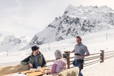 Ein Kellner serviert zwei Gästen auf einer Sonnenterrasse im Skigebiet das Essen. Die Gäste sitzen an einem Holztisch mit Kaffee und genießen den Blick auf die schneebedeckten Berge im Hintergrund. Der Himmel ist klar und die Sonne beleuchtet die Winterlandschaft. | © Gargellner Bergbahnen GmbH & Co KG, Daniel Zangerl