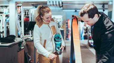 In einem Skiverleih berät ein Mitarbeiter eine Frau zur passenden Tourenausrüstung. Die Frau hält einen türkisfarbenen Skitourenschuh der Marke Scarpa, während der Mitarbeiter ein Paar Ski mit orangefarbenen Fellen zeigt. Im Hintergrund sind weitere Ski und Ausrüstungsgegenstände zu sehen. | © Gargellner Bergbahnen GmbH & Co KG, Christoph Schöch