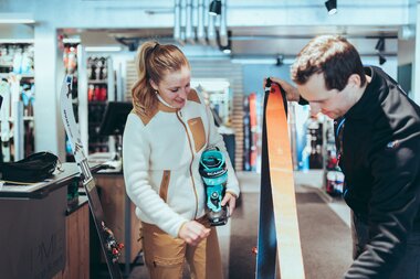 In einem Skiverleih berät ein Mitarbeiter eine Frau zur passenden Tourenausrüstung. Die Frau hält einen türkisfarbenen Skitourenschuh der Marke Scarpa, während der Mitarbeiter ein Paar Ski mit orangefarbenen Fellen zeigt. Im Hintergrund sind weitere Ski und Ausrüstungsgegenstände zu sehen. | © Gargellner Bergbahnen GmbH & Co KG, Christoph Schöch