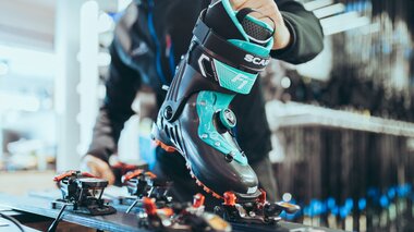 Eine Person passt in einer Werkstatt einen Skitourenschuh der Marke Scarpa an die Bindung eines Tourenskis an. Der schwarze Schuh mit türkisfarbenen Akzenten wird auf die vordere Bindung gesetzt. Im Hintergrund sind Regale mit Skiausrüstung zu erkennen. | © Gargellner Bergbahnen GmbH & Co KG, Christoph Schöch