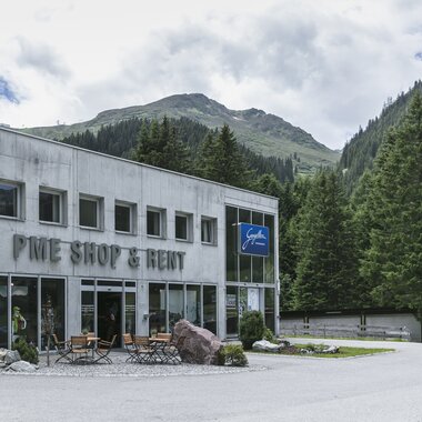 Das Gebäude des PME Shop & Rent in Gargellen steht vor einer Kulisse aus grünen Bergen und dichten Wäldern. Das zweistöckige Haus aus grauem Beton hat große Glasfronten im Erdgeschoss mit der Aufschrift „PME Shop & Rent“ sowie dem blauen Schild „Gargellen Montafon“. Vor dem Eingang stehen Tische und Stühle auf einer kleinen Terrasse. | © Gargellner Bergbahnen GmbH & Co KG, Christoph Schöch