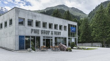 Das Gebäude des PME Shop & Rent in Gargellen steht vor einer Kulisse aus grünen Bergen und dichten Wäldern. Das zweistöckige Haus aus grauem Beton hat große Glasfronten im Erdgeschoss mit der Aufschrift „PME Shop & Rent“ sowie dem blauen Schild „Gargellen Montafon“. Vor dem Eingang stehen Tische und Stühle auf einer kleinen Terrasse. | © Gargellner Bergbahnen GmbH & Co KG, Christoph Schöch