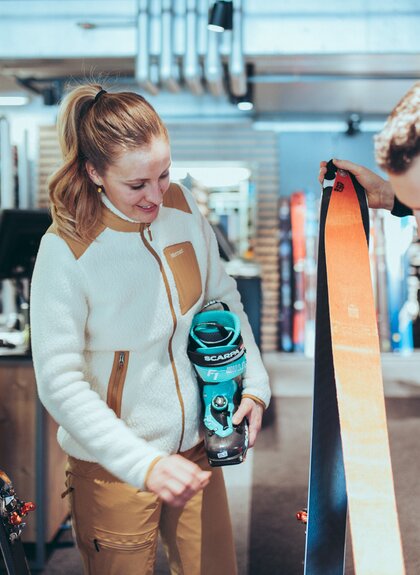 In einem Skiverleih berät ein Mitarbeiter eine Frau zur passenden Tourenausrüstung. Die Frau hält einen türkisfarbenen Skitourenschuh der Marke Scarpa, während der Mitarbeiter ein Paar Ski mit orangefarbenen Fellen zeigt. Im Hintergrund sind weitere Ski und Ausrüstungsgegenstände zu sehen. | © Gargellner Bergbahnen GmbH & Co KG, Christoph Schöch