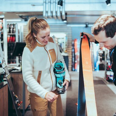 In einem Skiverleih berät ein Mitarbeiter eine Frau zur passenden Tourenausrüstung. Die Frau hält einen türkisfarbenen Skitourenschuh der Marke Scarpa, während der Mitarbeiter ein Paar Ski mit orangefarbenen Fellen zeigt. Im Hintergrund sind weitere Ski und Ausrüstungsgegenstände zu sehen. | © Gargellner Bergbahnen GmbH & Co KG, Christoph Schöch