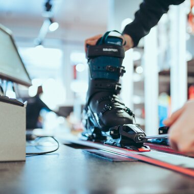 Eine Person stellt mit einem Schraubenzieher die Bindung eines Skis ein, in dem ein schwarzer Skischuh der Marke Salomon eingespannt ist. Der Ski liegt auf einer Werkbank in einem modernen Skiverleih. Im Hintergrund sind Arbeitsgeräte und Regale leicht unscharf zu erkennen. | © Gargellner Bergbahnen GmbH & Co KG, Christoph Schöch