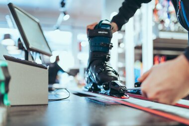 Eine Person stellt mit einem Schraubenzieher die Bindung eines Skis ein, in dem ein schwarzer Skischuh der Marke Salomon eingespannt ist. Der Ski liegt auf einer Werkbank in einem modernen Skiverleih. Im Hintergrund sind Arbeitsgeräte und Regale leicht unscharf zu erkennen. | © Gargellner Bergbahnen GmbH & Co KG, Christoph Schöch