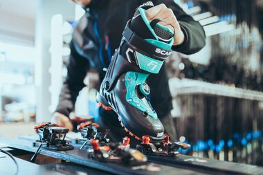 Eine Person passt in einer Werkstatt einen Skitourenschuh der Marke Scarpa an die Bindung eines Tourenskis an. Der schwarze Schuh mit türkisfarbenen Akzenten wird auf die vordere Bindung gesetzt. Im Hintergrund sind Regale mit Skiausrüstung zu erkennen. | © Gargellner Bergbahnen GmbH & Co KG, Christoph Schöch