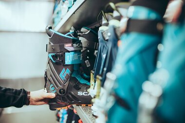 Eine Person nimmt einen schwarzen und türkisfarbenen Tourenskischuh aus einem Regal mit mehreren Paaren. Der Schuh trägt die Aufschrift „Scarpa F1“. Die Aufnahme zeigt den Moment der Auswahl in einem Skiverleih oder Sportgeschäft. | © Gargellner Bergbahnen GmbH & Co KG, Christoph Schöch