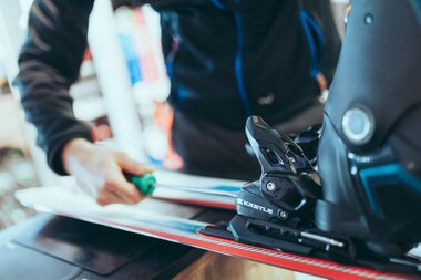 Eine Person justiert mit einem Schraubenzieher die Bindung eines Skis der Marke Kästle. Der Vorderteil des Skischuhs steckt in der Bindung, während im Hintergrund Werkstattausrüstung leicht unscharf zu sehen ist. Die Szene zeigt den Servicevorgang in einer Skiverleih- oder Werkstattumgebung. | © Gargellner Bergbahnen GmbH & Co KG, Christoph Schöch