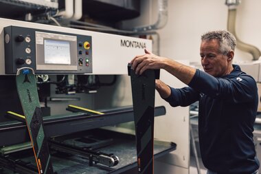 Ein Mitarbeiter des PME Shop & Rent steht in der Werkstatt an einer Skiservicemaschine und hält einen Ski in den Händen; neben ihm ist ein weiterer Ski eingespannt, die Maschine mit Bedienfeld ist im Hintergrund sichtbar. | © Gargellner Bergbahnen GmbH & Co KG, Daniel Zangerl