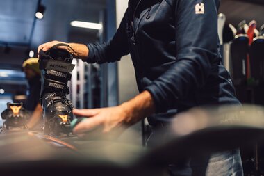 Ein Mitarbeiter des PME Shop & Rent arbeitet im Verleihbereich an einem Skischuh, der in eine Skibindung eingesetzt ist; der Skischuh wird mit beiden Händen justiert, im Hintergrund sind weitere Skier zu erkennen. | © Gargellner Bergbahnen GmbH & Co KG, Daniel Zangerl