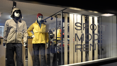 Blick durch das Schaufenster eines Sport- und Modegeschäfts. Zwei Schaufensterpuppen tragen Outdoorjacken und Skihosen, eine in Beige, die andere in Gelb mit roter Mütze und Sonnenbrille. Im Hintergrund sind Regale mit weiterer Skibekleidung zu sehen. Auf der Scheibe steht groß der Schriftzug „SPORT MODE RENT“. | © Gargellner Bergbahnen GmbH & Co KG