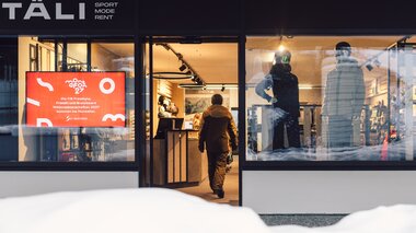 Außenansicht des Sportshops TÄLI Sport Mode Rent in Gargellen; eine Person betritt den beleuchteten Shop durch den Eingang, Schaufenster mit Sportbekleidung und Beschilderung sind sichtbar, Schnee liegt vor dem Gebäude. | © Gargellner Bergbahnen GmbH & Co KG, Daniel Zangerl