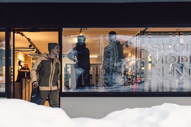 Außenansicht des Sportshops TÄLI Sport Mode Rent in Gargellen; ein Mann verlässt den Shop mit einer Einkaufstasche, im Schaufenster sind Schaufensterpuppen mit Wintersportbekleidung zu sehen, Schnee liegt vor dem Gebäude. | © Gargellner Bergbahnen GmbH & Co KG, Daniel Zangerl